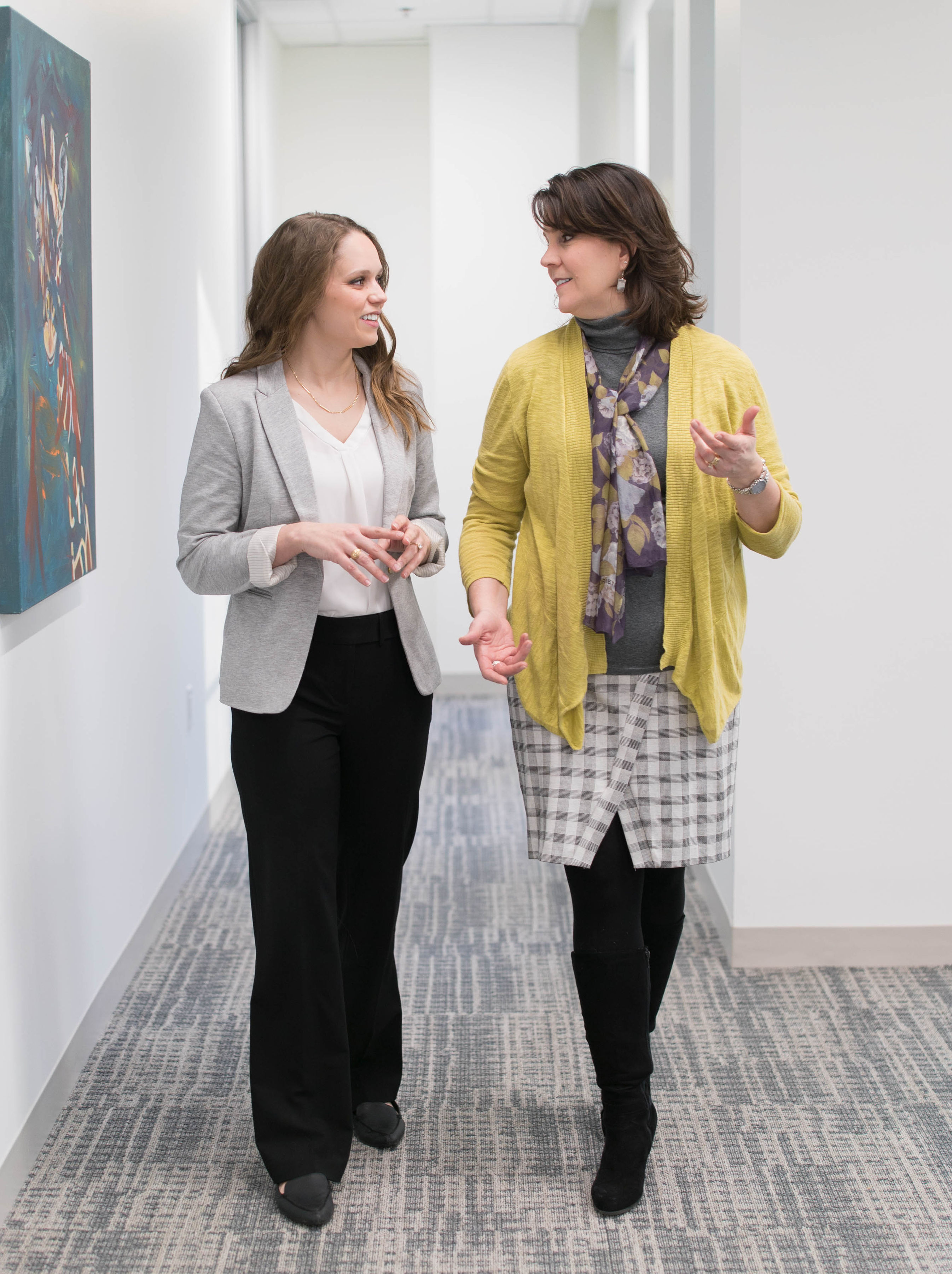 Two Caucasian females walking down a hallway conversing with one another while using their hands to speak
