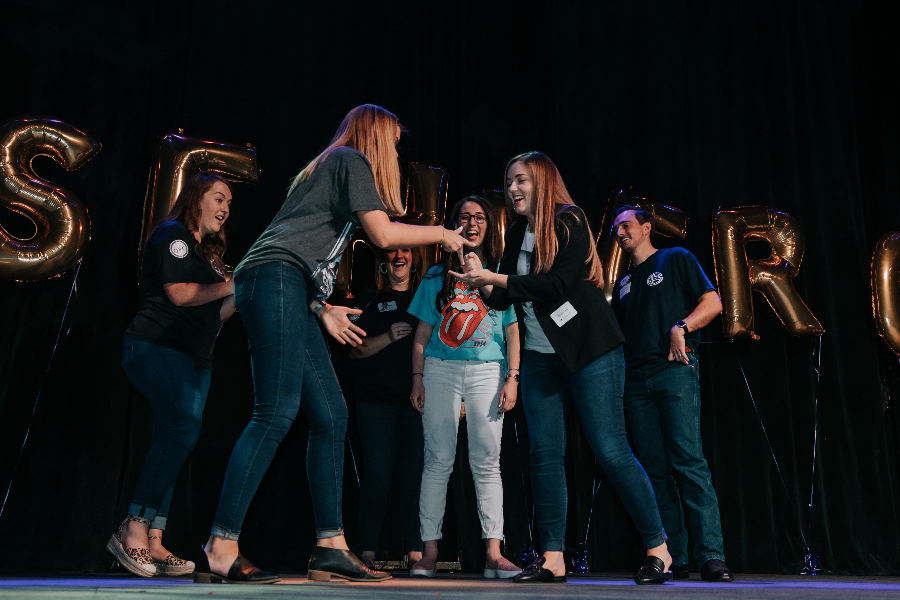 Two Caucasian females participating in a rock, paper, scissors competition on stage at an all-company meeting