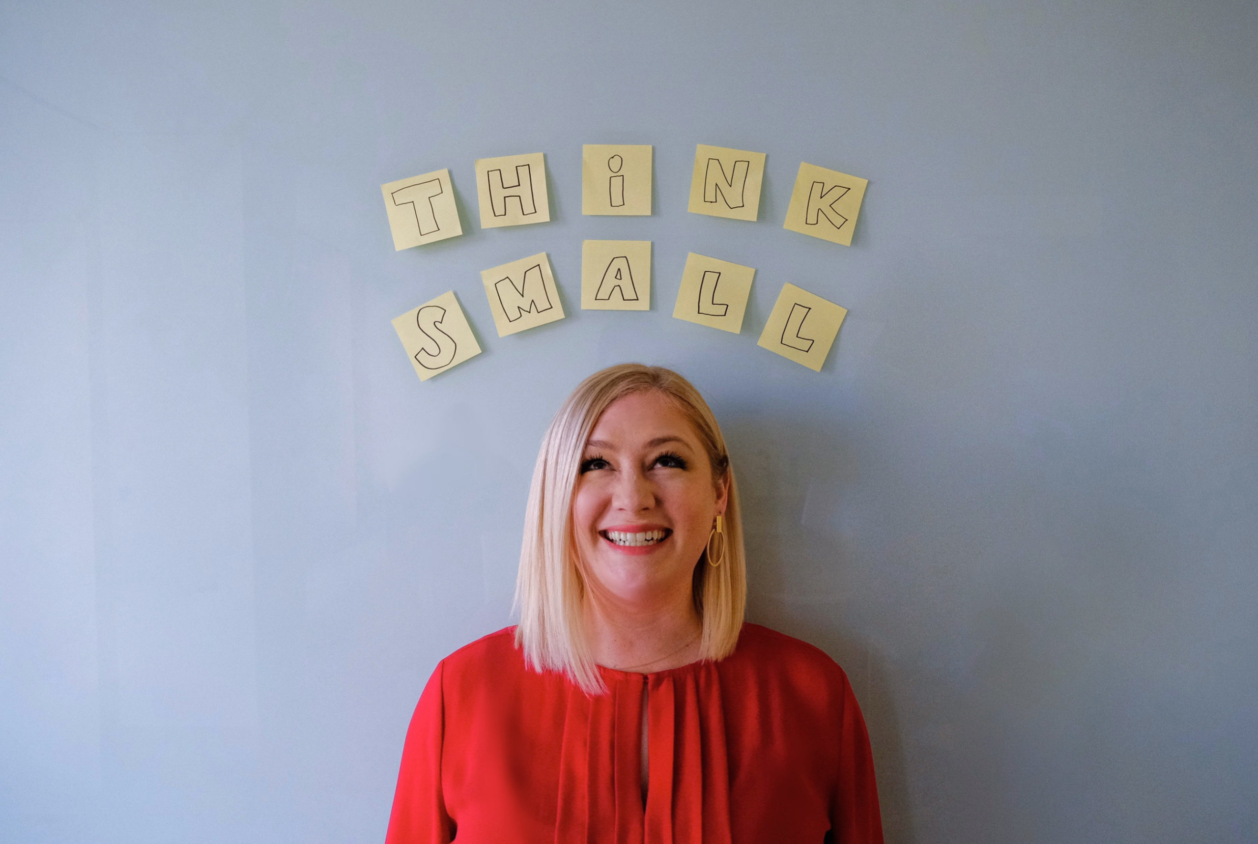 A Caucasian female in a red blouse looking at sticky notes that read, "Think Small" above her head that are attached to a wall
